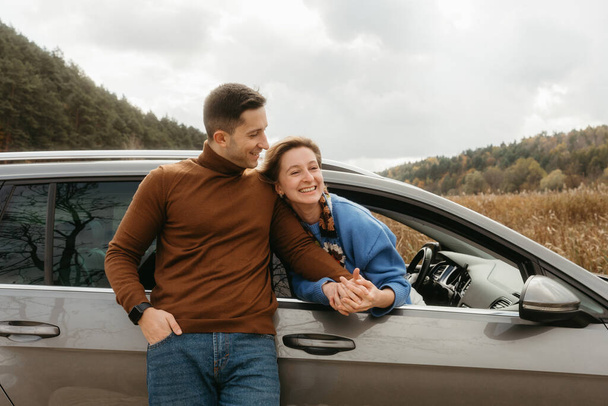Uomo felice appoggiato alla macchina e che tiene per mano la donna allegra che seduta all'interno, coppia adulta godendo di viaggio su strada in autunno - Foto, immagini