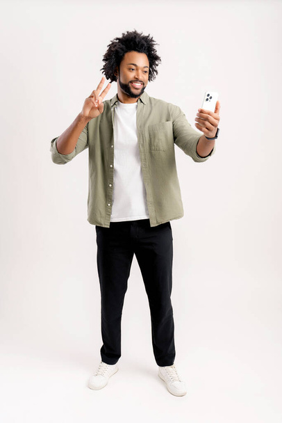 Full length view of the young man showing victory gesture with his hand while chatting via cell phone or making selfie, isolated on white background - Photo, Image