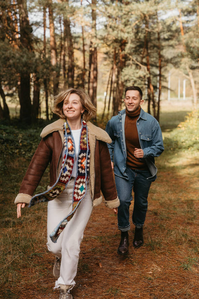 Mutlu çift ormanın ortasında eğleniyor, gülen kadın kocasıyla birlikte sonbahar parkında koşuyor. - Fotoğraf, Görsel