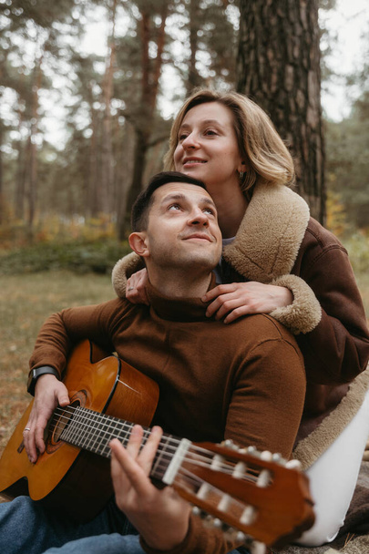 Egy férfi gitározik, és a nő hátulról öleli meg az őszi parkban. - Fotó, kép