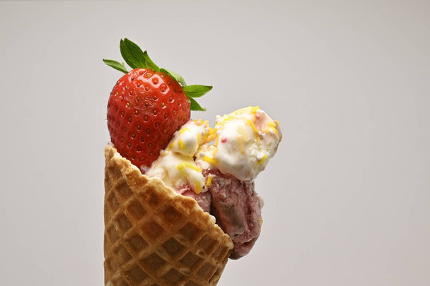 Délicieuses boules de glace à la vanille et aux baies dans un cône de gaufre garni d'une fraise fraîche et d'une feuille de menthe sur fond blanc - Photo, image