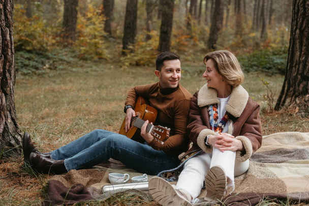 Egy vidám férfi gitározik egy boldog nőnek, miközben ők a takarón ülnek az őszi erdőben. - Fotó, kép