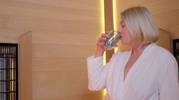 Portrait de belle femme blonde buvant de l'eau dans un salon de spa. Une femme en peignoir pendant les soins spa - Séquence, vidéo
