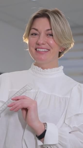 Μια ξανθιά γυναίκα με χαρούμενα μάτια φοράει ένα λευκό πουκάμισο με τυλιγμένα μανίκια, τα μαλλιά της πέφτουν με χάρη πάνω από τους ώμους της καθώς χαμογελά σε μια ευχάριστη χειρονομία σε μια εκδήλωση. - Πλάνα, βίντεο