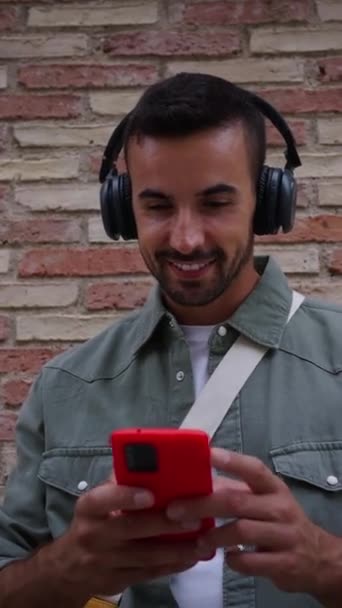 Вертикально привлекательный молодой человек с наушниками, использующий красный сотовый телефон, опирающийся на кирпичную стену. Тысячелетний европейский мужчина наслаждается взглядом, улыбаясь, стоя на улице. Люди, зависимые от технологий  - Кадры, видео
