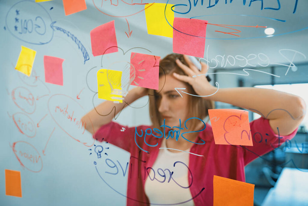 Portrait of young caucasian businesswoman thinking with confused face expression while standing in front of glass board with sticky notes and mind map at creative business meeting. Immaculate. - Photo, Image