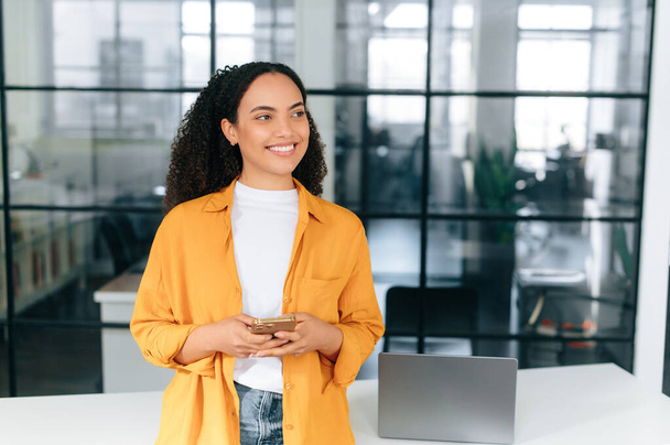 Stilvolle positive brasilianische oder hispanische lockige junge Frau in orangefarbenem Hemd, steht an einem Arbeitsplatz im Büro, hält ihr Smartphone in der Hand, sendet Nachrichten online, schaut weg, lächelt, träumt - Foto, Bild