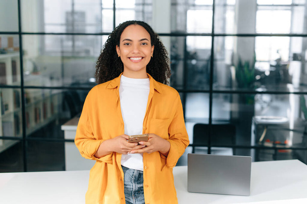Happy beautiful brazilian or hispanic curly haired young woman in an orange shirt, stands at a workplace in the office, holding her mobile phone, chatting online, looks at camera, smiling friendly - 写真・画像