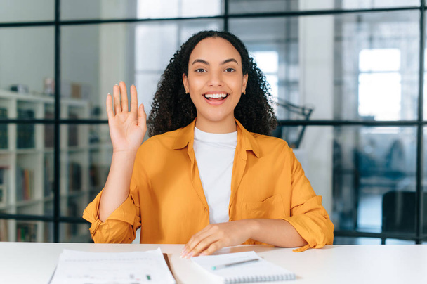 Positive hispanic or brazilian curly haired young woman, communicates via video conference, listens or conducts an online lesson, coaching session, lecture, brainstorm, makes a greeting sign, smiles - Photo, Image