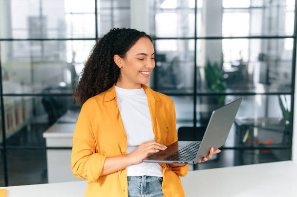 Umtriebige positive brasilianische oder hispanische lockige Frau in lässiger Kleidung, Firmenmanagerin, seo, mit offenem Laptop, steht in einem modernen Kreativbüro, schaut auf den Bildschirm, lächelt - Foto, Bild