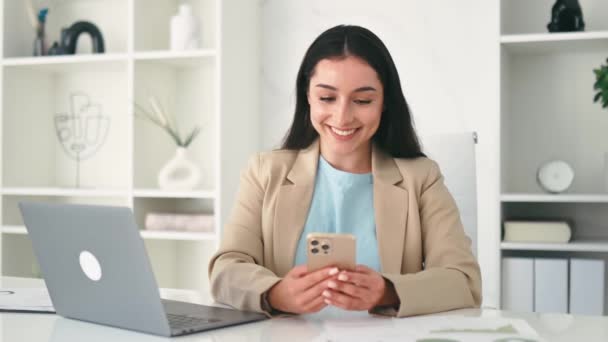 Позитивна арабська або індійська жінка, одягнена в елегантний одяг, співробітник компанії, секретар, сидить на робочому місці в офісі, використовує свій смартфон, обмін повідомленнями з друзями або клієнтами, дивиться на екран, посміхається - Кадри, відео