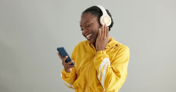Black woman, headphones and smartphone for music with dancing in studio, fun and audio streaming on grey background. Listening to radio playlist, tech and energy with happiness, mobile app and rhythm. - Footage, Video