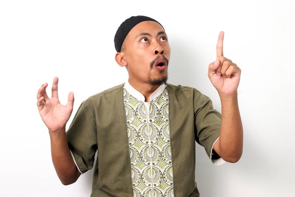 Ενθουσιασμένος Ινδονήσιος Μουσουλμάνος με πουκάμισο Κόκο και πέτσι έχει μια ιδέα, να υψώσει το δάχτυλό του σε μια στιγμή εύρηκα. Απομονωμένα σε λευκό φόντο - Φωτογραφία, εικόνα