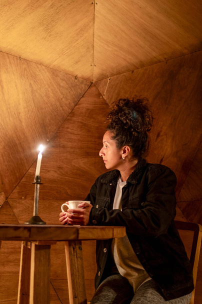 Χαλαρή γυναίκα που πίνει ένα φλιτζάνι καφέ καθισμένη μέσα σε μια σκηνή θόλου φωτισμένη με κεριά. - Φωτογραφία, εικόνα