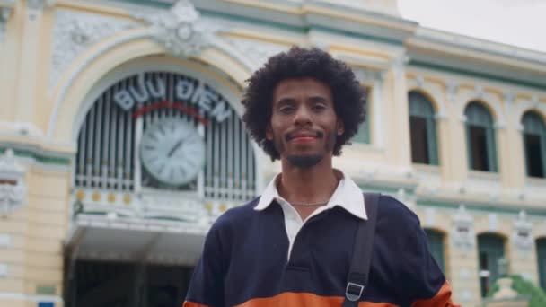 Niski kąt portret czarnego wesołego człowieka uśmiechającego się do kamery stojącej na tle budynku miasta - Materiał filmowy, wideo