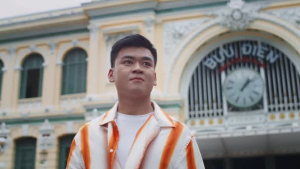Şehir merkezinde kameraya gülümseyen çizgili gömlekli neşeli Asyalı genç adamın alçak açılı portresi. - Video, Çekim