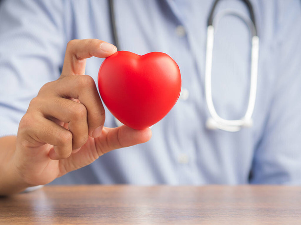 Mão de um médico mostrando uma forma de coração vermelho enquanto sentado no hospital ou clínica. Fotografia de perto. Espaço para SMS. Conceito médico e de incentivo. - Foto, Imagem