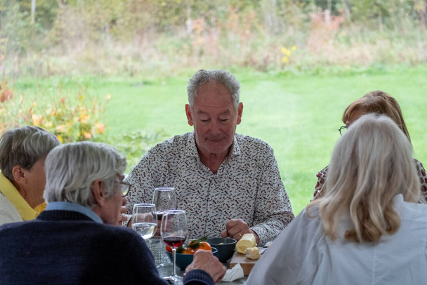Dit beeld toont een oudere man die geniet van een maaltijd met vrienden. Hij glimlacht en lijkt zich bezig te houden met een aangenaam gesprek met de vrouw tegenover hem. Ze zitten aan een tafel beladen met - Foto, afbeelding
