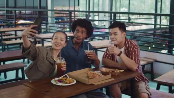 Medium lange shot van drie vrolijke vrienden nemen selfie op smartphone met drankjes en pizza zitten in food-court - Video