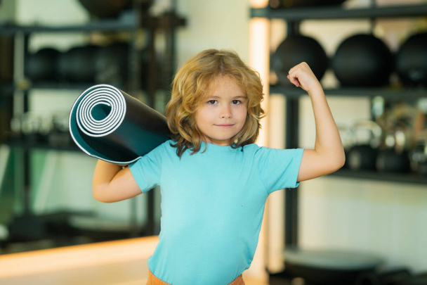 Excited kid with strong arm gesture holding yoga mat in gym. Yoga child concept. Young strong sporty kid. Workout sport concept. - Photo, image