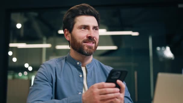 Close-up van volwassen man met baard met behulp van moderne smartphone en glimlachen tijdens het zitten op coworking ruimte. Tevreden mannelijke manager krijgt een geweldig idee tijdens het surfen op internet en na te denken over het project. - Video