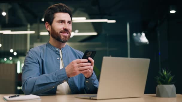 Mann im schicken Hemd tippt auf Smartphone und lächelt, während er am Schreibtisch mit Computer sitzt. Männlicher Angestellter benutzt modernes Gerät zur Unterhaltung und chattet mit Freund, während er Pause von der Arbeit hat. - Filmmaterial, Video
