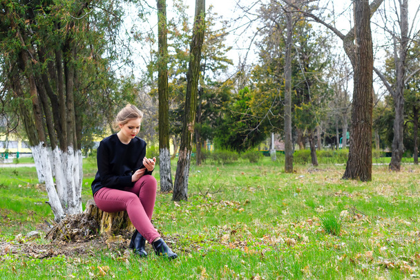 Beautiful girl sitting on a stump in the park with your smartpho - Foto, Imagen