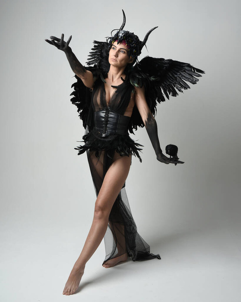 Full length portrait of female model wearing gothic horned headdress with halloween black dress and fantasy angel feather wings. Standing walking, holding pose bird prop. Isolated studio background - Photo, Image