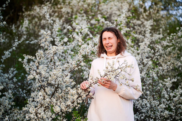 Femme allergique souffrant d'allergie saisonnière au printemps, posant dans le jardin en fleurs au printemps. Portrait de jeune femme les yeux fermés éternuant parmi les arbres en fleurs. Concept d'allergie printanière - Photo, image