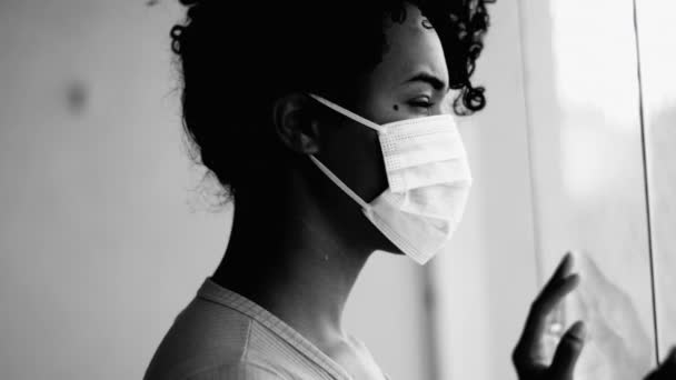 Yksi lukittu nuori musta nainen vangittiin kotona pandemian aikana katselee ulos huoneiston ikkunasta yllään kirurginen kasvonaamio tunne loukussa, yksivärinen - Materiaali, video