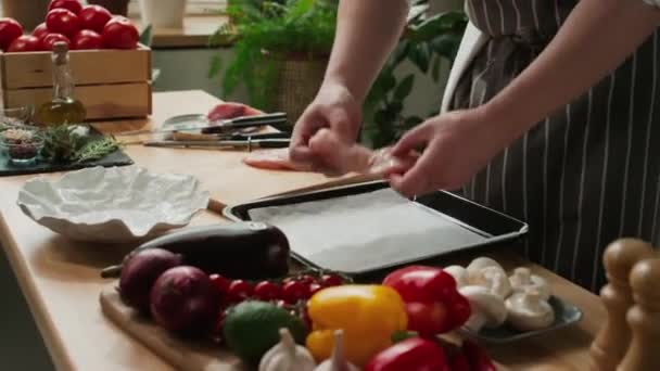 Обрізаний знімок невизначеного шеф-кухаря-кухаря в смугастому фартусі, що розміщує дві сирі курячі грудки на пергаменті на сковороді - Кадри, відео