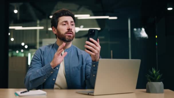 Houkutteleva mies puhuu ja katselee älypuhelimen näyttöä videopuhelun aikana. Mies freelancer istuu pöydän ääressä kannettavan tietokoneen kanssa ja kommunikoi työsopimuksesta online-konferenssin aikana. - Materiaali, video