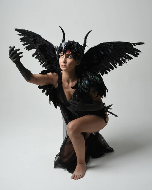 Portrait complet de mannequin femme portant une coiffure gothique à cornes avec robe noire d'Halloween et ailes fantaisistes de plumes d'ange. Pose accroupie, agenouillée sur le sol. Fond de studio isolé - Photo, image