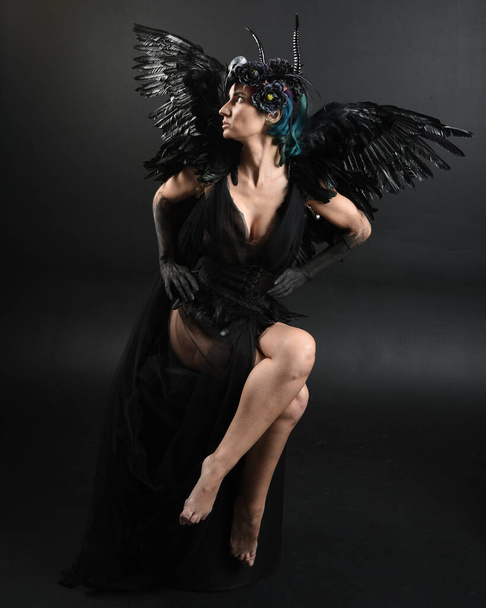 Full length πορτρέτο του μοντέλου φορώντας gothic κέρατο φόρεμα κεφαλής με αποκριές μαύρο φόρεμα, φτερά φτερό αγγέλου φαντασίας. Σκυμμένη πόζα. Απομονωμένο σκοτεινό φόντο στούντιο, κινηματογραφικές σκιές σιλουέτες  - Φωτογραφία, εικόνα
