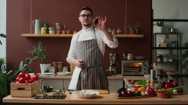 Scatto medio di cuoco maschio caucasico in abiti da lavoro e occhiali in piedi vicino al tavolo da cucina in legno in cucina e dimostrando come tagliare avocado verde maturo alla fotocamera - Filmati, video