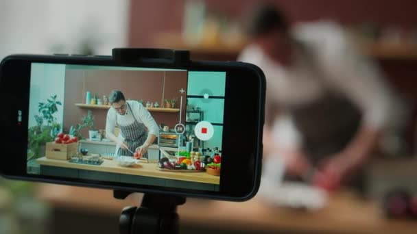 Prostřednictvím displeje smartphonu záběr kavkazské mladý muž kuchař v pracovním oblečení natáčení vaření videa v kuchyni, demonstrující, jak krájet grapefruit s nožem na dřevěné desce - Záběry, video