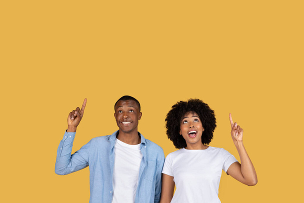 Optimistic shocked millennial African American couple looking up and pointing towards the sky with hopeful expressions, standing against a plain yellow background, studio - Photo, Image
