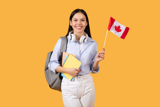 Fröhliche Studentin mit strahlendem Lächeln hält kanadische Flagge und Notizbücher mit Kopfhörern um den Hals und symbolisiert akademischen Enthusiasmus und Nationalstolz vor leuchtend gelbem Hintergrund - Foto, Bild