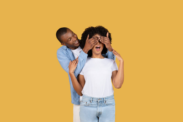 Een speelse duizendjarige Afro-Amerikaanse man verrast een vrouw door haar ogen van achteren te bedekken, zowel lachend als staand tegen een effen gele achtergrond, studio. Romantische date emoties - Foto, afbeelding