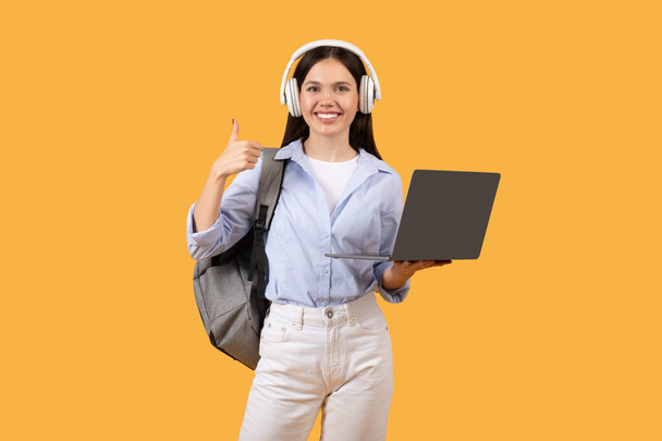 Fröhliche Studentin reicht Daumen hoch, während sie Laptop und Kopfhörer um den Hals hält, was Erfolg und Anerkennung in einer E-Learning-Umgebung vor gelbem Hintergrund symbolisiert - Foto, Bild