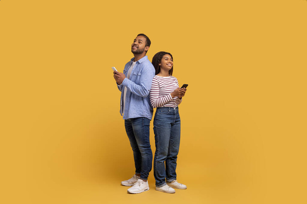 背中に立っている若い黒いカップルは,それぞれスマートフォンに魅了され,現代の接続を描いた幸せなアフリカ系アメリカ人男性と女性,活気に満ちた黄色の背景にポーズ - 写真・画像