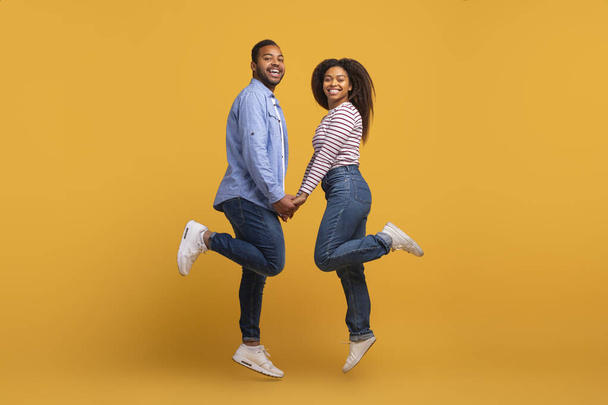 Happy young black couple holding hands and jumping in unison, african american man and woman showcasing happiness and togetherness, having fun against warm yellow studio background, copy space - Photo, Image