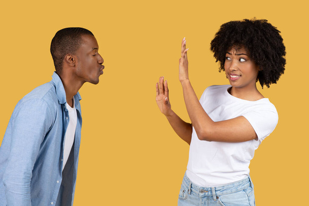 Man blowing a kiss to a millennial woman who playfully rejects it with a hand gesture, both dressed in casual clothing against a yellow background, studio. Say no and stop harassment - Foto, afbeelding