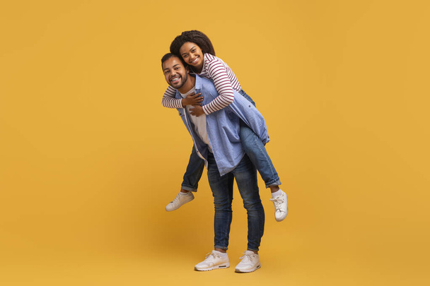 Delighted young black couple having fun together against yellow background in studio, cheerful african american man giving piggyback ride to his happy woman, smiling joyfully at camera, copy space - Photo, Image