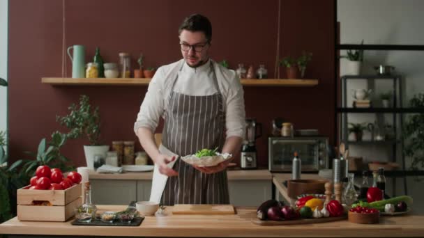 Medium shot of young Caucasian male chef with microphone describing and showing process of salad preparation at camera, sprinkling seeds on it while finishing - Footage, Video