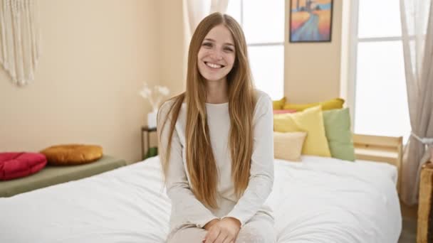 Fiatal, gyönyörű szőke nő pizsamában ül boldogan az ágyon, hűvös mosolya kivilágítja a szobát a kényelmes lakásában. - Felvétel, videó