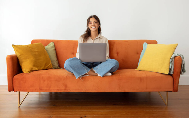Портрет красивої усміхненої молодої жінки з ноутбуком на лапах сидячи на перехресті на дивані вдома, приваблива міленіалізована леді працює або навчається онлайн, позує з комп'ютером у вітальні Інтер'єр - Фото, зображення