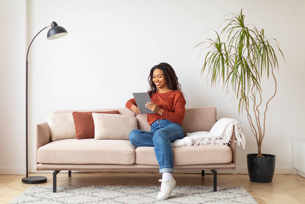 Красива молода чорна жінка, використовуючи цифровий планшет вдома, посміхається афроамериканська жінка відпочиває на дивані у вітальні з сучасним гаджетом, переглядаючи Інтернет або покупки в Інтернеті, копіювати простір - Фото, зображення