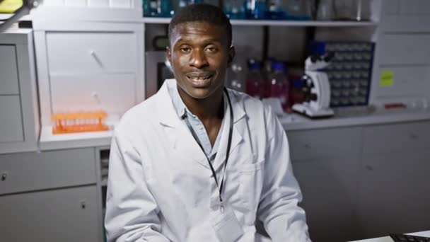 Alegre científico afroamericano masculino con guantes apunta con confianza y sonríe a algo al costado en el entorno del laboratorio - Metraje, vídeo