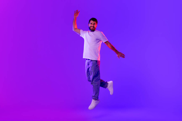 Joyful carefree young man jumps and raises arm posing in mid air, in blue and purple neon lights over studio background, smiling to camera. Concept of happiness, good news celebration - Photo, image